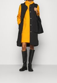 Schwarze, gesteppten lange Weste mit goldenen Knöpfen, getragen über einem gelben Hoodie-Kleid, kombiniert mit kniehohen schwarzen Stiefeln. Einfaches, funktionales Design.