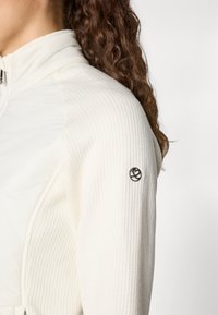 Weißer Blazer mit gerippter Textur und glatten Paneelen, versehen mit einem schwarzen Logo auf dem linken Ärmel. Mit Reißverschluss vorne und hohem Kragen.