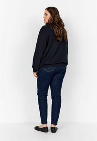 Sudadera negra con un corte relajado, combinada con jeans ajustados de color azul oscuro y bailarinas negras trenzadas. El cabello es largo y ondulado. Detalles mínimos en el diseño.