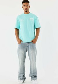 Camiseta de algodón azul claro de mangas cortas con estampado frontal, combinada con jeans azules deslavados que presentan detalles de patchwork y doble costura.