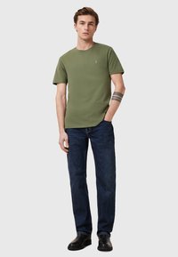 Oliivinvihreä lyhythihainen t-paita pyöreällä kaula-aukolla ja pienellä logolla, yhdistettynä tummansinisiin suoriin farkkuihin ja mustiin saappaisiin.