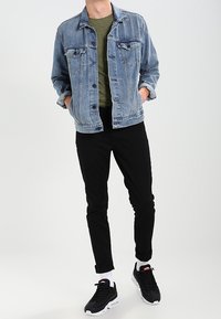 Džínová bunda v světle modré, zelené tričko, černé slim-fit kalhoty a černé tenisky s bílou podešví a červenými akcenty. Jednoduché pozadí.