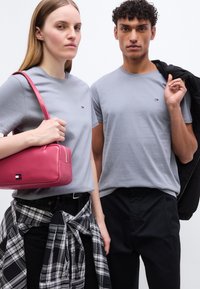 Δύο μοντέλα φοράνε ανοιχτό γκρι μπλουζάκια· η γυναίκα κρατάει ροζ τσάντα και έχει πουκάμισο καρό δεμένο γύρω από τη μέση, ο άντρας κρατάει μαύρο σακάκι πάνω στον ώμο.