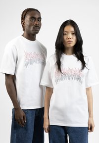 White cotton t-shirts feature graphic designs in red and black, with a relaxed fit. Paired with denim shorts, showcasing a casual style.