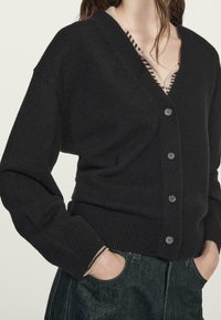 Czarny dziergany kardigan z dekoltem w serek, zapinany na pięć guzików, z prążkowanymi mankietami i luźnym krojem, noszony na paskowanym topie.