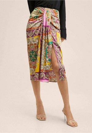 Knee-length multicolored patterned skirt with side slit worn with black top and transparent high-heeled sandals on neutral background.
