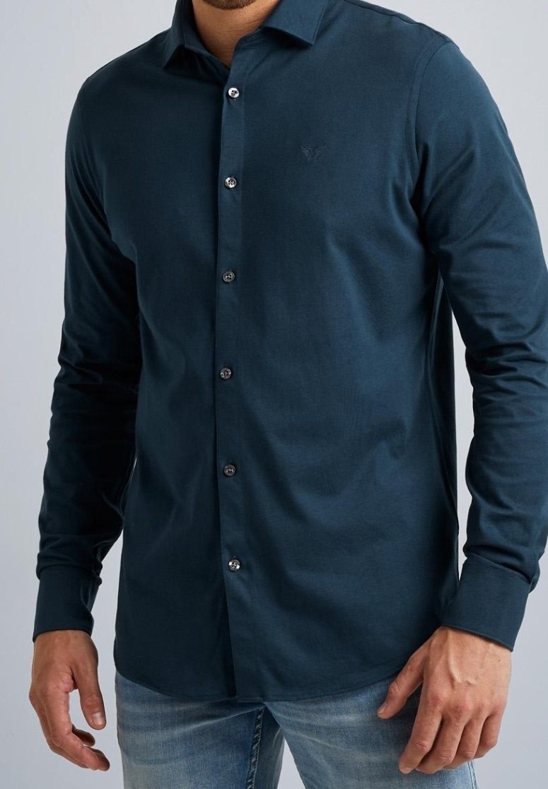 Homme portant une chemise bleu foncé à manches longues boutonnée avec un jean bleu clair, mains détendues sur les côtés, sur un fond uni.