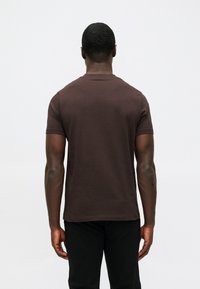 Brūna īsām piedurknēm t-kreklu ar apaļu kakla izgriezumu un gludu tekstūru, valkā ar melniem biksēm. Aizmugures skats rāda pieguļošu siluetu.