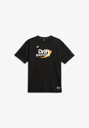 Młody mężczyzna o krótkich włosach nosi czarną koszulkę Drift Masters i czarne spodnie, stoi na jednolitym szarym tle.