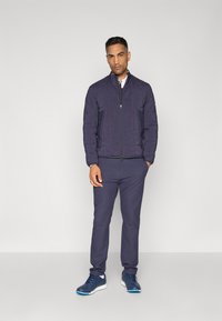 Veste de bombardier marine avec col montant et deux poches latérales zippées, associée à un pantalon marine et des chaussures de sport bleues sur un fond gris.