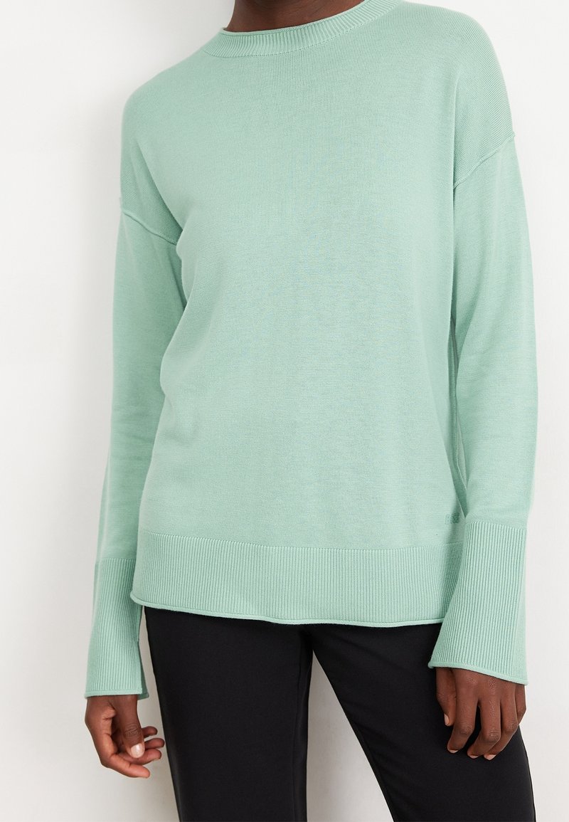 Personne portant un pull vert menthe à manches longues avec poignets et ourlet côtelés, associé à un pantalon noir sur un fond clair uni.
