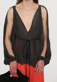 Blouse noire plissée avec un décolleté en V profond, manches fluides, attachée à la taille. Associée à une jupe orange vibrante et un sac à main texturé.