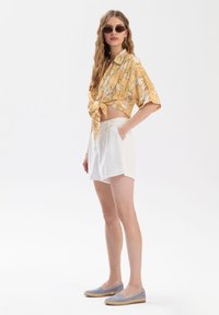 Żółta, kwiecista koszula z krótkim rękawem zawiązana w pasie, białe lniane szorty oraz szare płócienne espadryle z jutowymi podeszwami. Brązowe okulary przeciwsłoneczne dopełniają stylizację.