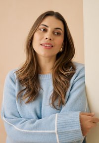 Pull en laine bleu clair avec poignets côtelés, surpiqûres blanches en accent. Texture douce, modèle aux cheveux longs appuyé contre un mur.