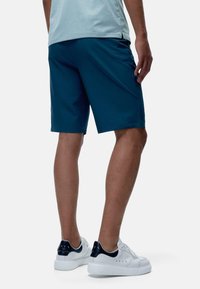 Jūras zila sporta šorti ar gludu tekstūru, iekšējām kabatām un brīvu piegriezumu, kombinēti ar baltiem apaviem ar jūras zilām akcentiem.