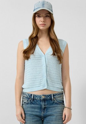 Junge Frau, die eine hellblaue Häkelweste mit Knöpfen, ausgewaschene blaue Jeans, eine Jeanskappe und ein silbernes Armband am rechten Handgelenk trägt.