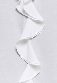 Tissu blanc présentant un détail de volants ondulés sur le bord, avec une texture lisse et un éclat subtil, posé sur un fond uni.