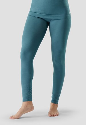 Close-up van een persoon die teal strakke leggings en een bijpassend topje draagt, op blote voeten staat met één voet licht verhoogd tegen een effen achtergrond.