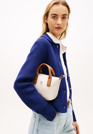 Femme portant un cardigan en maille bleu et un jean clair, tenant un petit sac à main blanc avec des poignées marron sous le bras.
