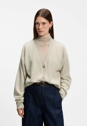 Frau mit glatten braunen Haaren trägt eine beigen Strickjacke über einem beigen Rollkragenpullover und dunkelblaue, hoch taillierte, weit geschnittene Jeans mit schwarzem Gürtel.