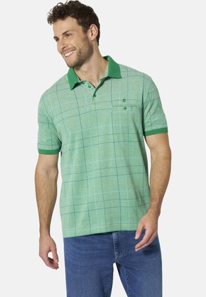 Lächelnder Mann trägt ein grünes kariertes Poloshirt mit kurzen Ärmeln, grünem Kragen und Ärmelsaum, kombiniert mit blauer Jeans.