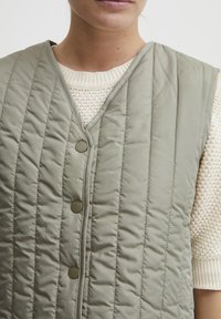 Gilet matelassé vert olive avec un col en V, doté de boutons et de coutures verticales. Porté sur un pull épais en tricot crème.