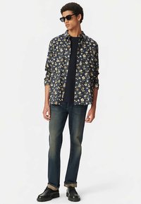 Marineblauw bloemenoverhemd met gele en witte patronen, gedragen over een zwart T-shirt, gecombineerd met een donkere spijkerbroek en zwarte schoenen.