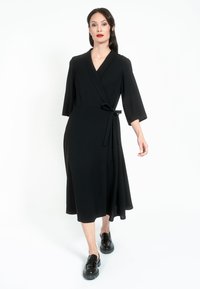 Fekete, V nyakú, széles ujjú ruha, derékon megkötővel. Simaságú anyag és térdhez érő szegély, fekete slip-on cipővel párosítva.