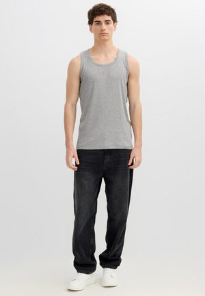 Joven de pie, vistiendo una camiseta sin mangas gris, vaqueros negros holgados y zapatillas blancas frente a un fondo claro y liso.