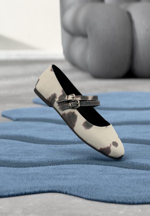 Beiger flacher Schuh mit schwarzen Flecken und doppelt geschnallten Riemen, der auf einem strukturierten blauen Teppich liegt.