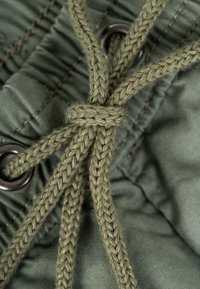 Close-up of green braided drawstring tied in a knot through metal eyelets on olive-green fabric with gathered stitching.