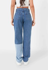 Mujer vestida con jeans azules bicolores y zapatillas blancas, vista desde atrás sobre un fondo claro y liso.