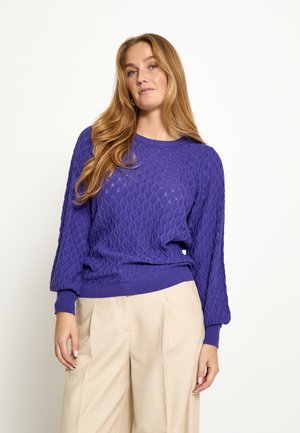 Naine, kellel on pikad blondid juuksed, kandmas tekstuuriga lilla pika varrukaga kampsunit ja beežid volditud pükse, taustaks tavaline valge taust.