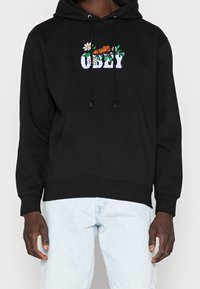 Svart hoodie i bomullstyg med en broderad design med blomsterdetaljer och ordet "UGLY" över bröstet.