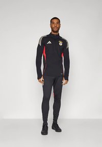 Vyras stovi vilkėdamas juodą ir raudoną Adidas Benfica treniruočių sportinį kostiumą su logotipais, priešais paprastą šviesią foną.