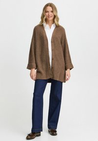 Brauner, gestrickter Cardigan mit strukturiertem Muster, großen Knöpfen und umgeschlagenen Ärmeln, kombiniert mit dunkelblauen, weiten Jeans und schwarzen Schuhen.