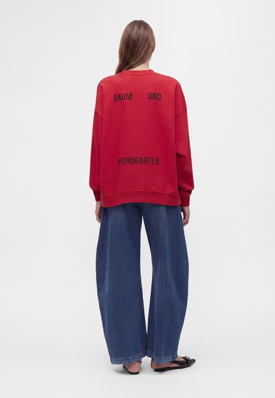 Personne aux cheveux longs porte un sweat-shirt rouge avec un texte noir et un jean large bleu, debout de dos sur un fond uni.