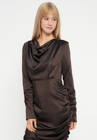 Rochie lungă cu mâneci lungi, de culoare maro închis, fabricată dintr-un material neted și lucios. Are un decolteu tip cowl și detalii drapate pe lateral.