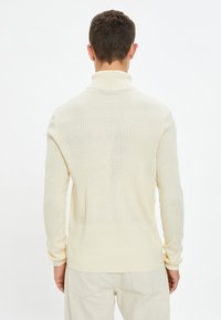 Cremfarbener gerippter Strickrollkragenpullover mit langen Ärmeln und taillierter Passform, aus einem strukturierten Stoff, getragen mit hellen Hosen.