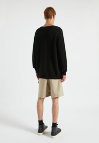 Personne aux cheveux courts portant un pull noir à manches longues, un short beige arrivant aux genoux, des chaussettes noires et des baskets montantes noires, debout et de dos.