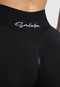 Zwarte high-waisted leggings met een textuurafwerking, voorzien van een wit geborduurd logo op de bovenband en versterkte naaddetails.