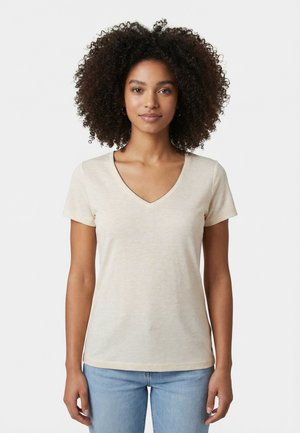 Giovane donna con capelli ricci che indossa una maglietta beige con scollo a V e maniche corte e jeans azzurri, in piedi davanti a uno sfondo bianco.