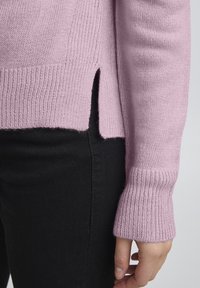Ljusrosa stickad tröja med ribbade muddar och sidofickor, kombinerad med svarta byxor, som visar upp en mjuk textur och avslappnad passform.