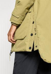 Olivengroene geïsoleerde jas met een ritssluiting aan de zijkant en knoopsluiting. Voorzien van een groot zakje aan de voorkant en manchetten met een gebreide textuur.