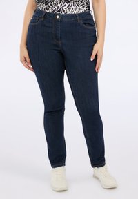 Vaqueros ajustados de denim azul oscuro con costuras naranjas, cinco bolsillos, cierre de botón y una textura suave. Llevados con zapatillas blancas.