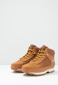 Botas de senderismo de cuero marrón con cordones amarillos y puños acolchados; cuenta con puntera reforzada y logo en el costado; suela de goma texturizada.