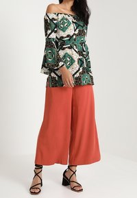 Donna che indossa una blusa a spalla scoperta con motivo, pantaloni larghi arancione ruggine e sandali neri con tacco a strappo, su sfondo neutro.