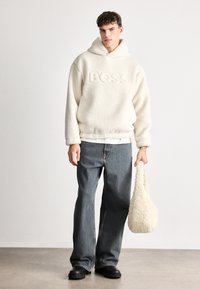 Kremeni dukserica od flisa s utisnutim natpisom "BOSS", u kombinaciji s plavim širokim farmerkama i crnim platformama, drži pahuljastu krem torbu.