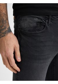 Pantalones vaqueros de mezclilla negra con un corte slim, que cuentan con bolsillos y un ligero desvanecimiento. La mano reposando en el costado resalta la textura y los detalles de la costura.