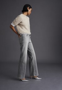Donna con capelli corti neri che indossa un maglione beige soffice, jeans grigio chiaro a gamba larga e tacchi alti argentati a punta, in piedi davanti a uno sfondo grigio.